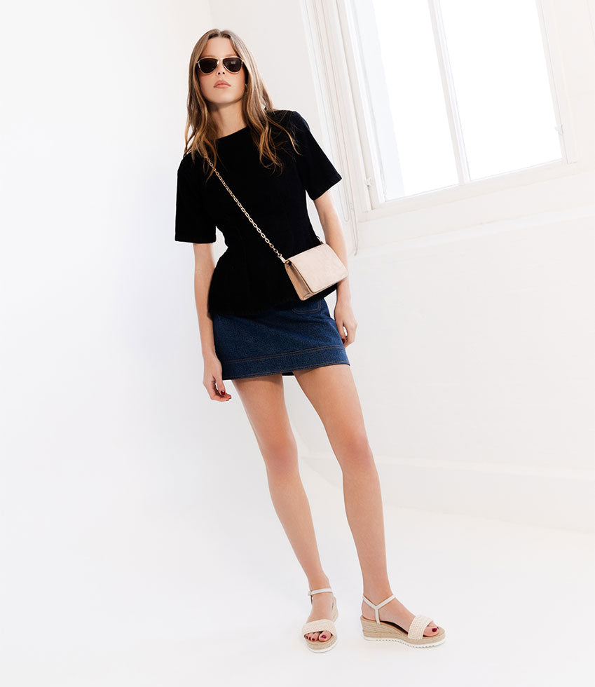 Woman wearing a black top and denim skirt with sunglasses and a beige bag on a white background