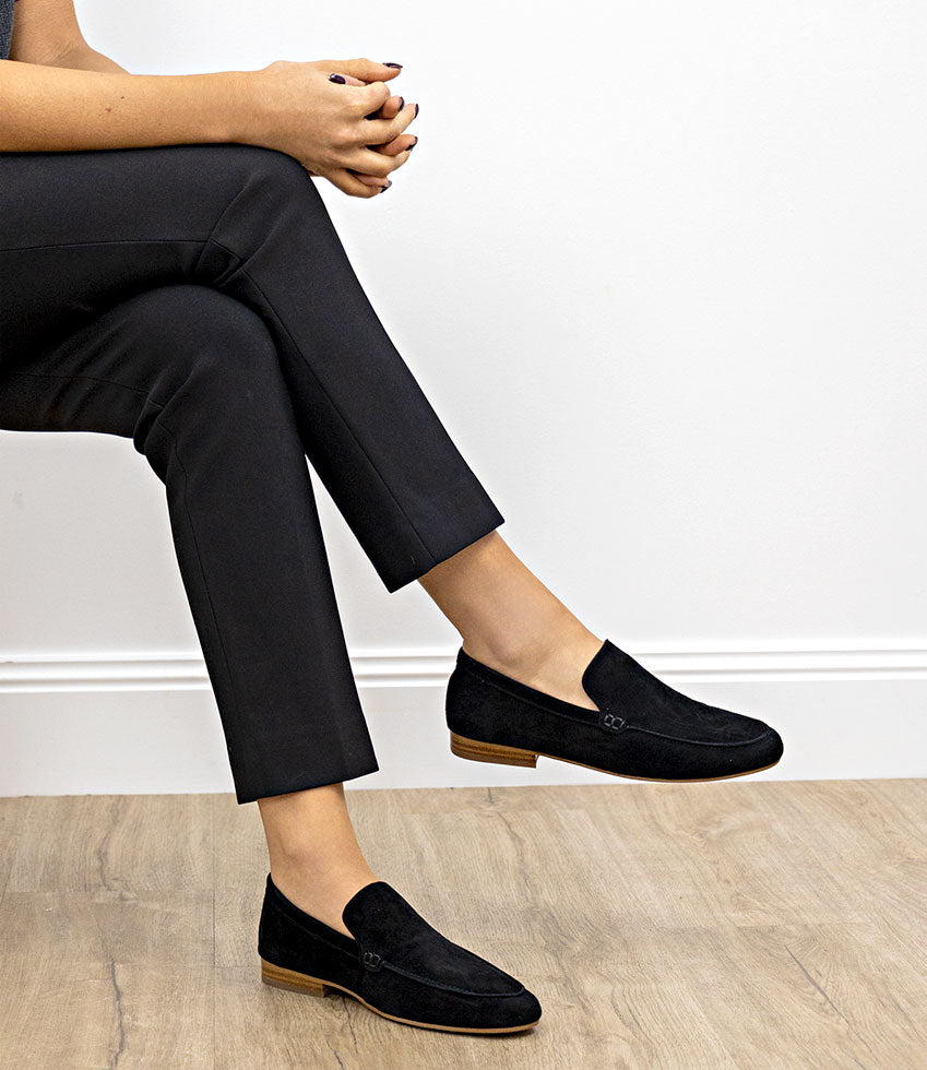 Person wearing HALT black suede loafers sitting on a wooden floor with a white wall background
