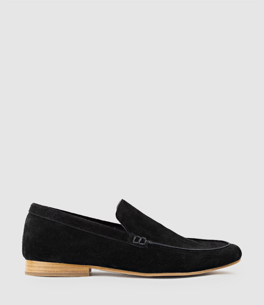 Person wearing HALT black suede loafers sitting on a wooden floor with a white wall background
